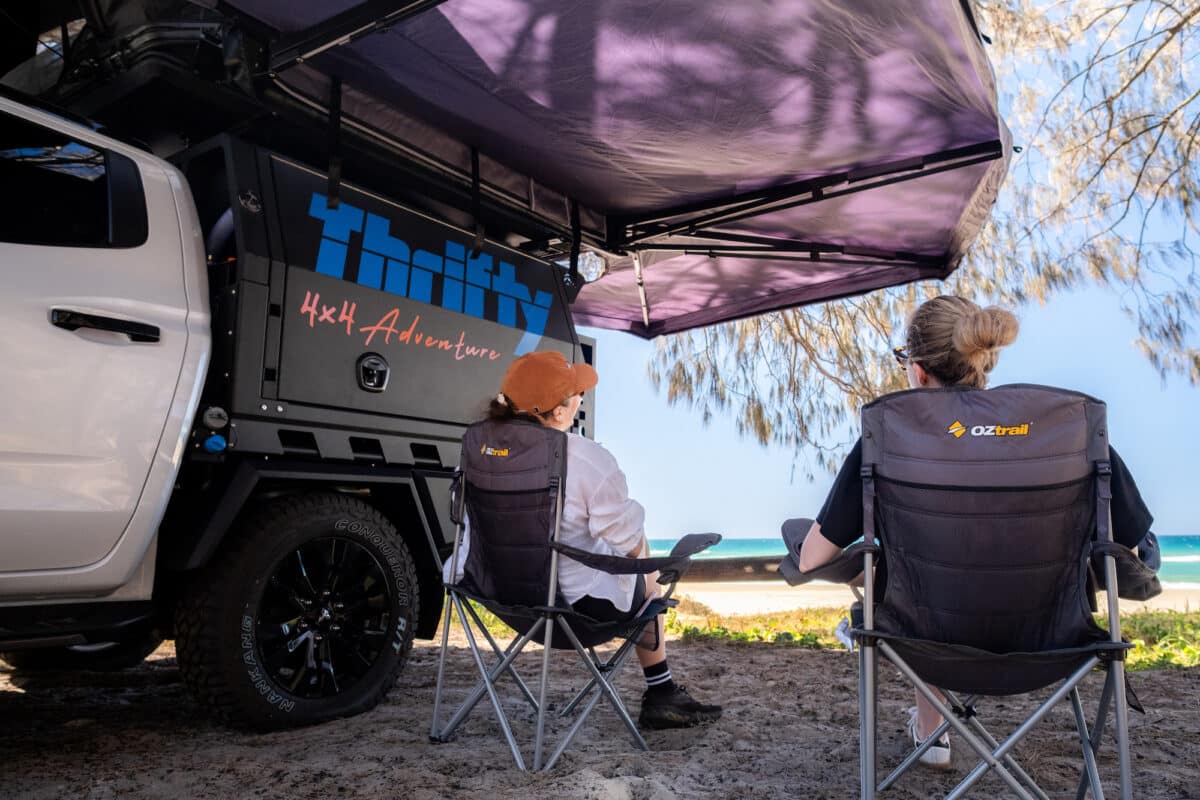 From behind: Two campers on campchairs sitting underneath a 270 degree awning fitted to a Thrifty 4X4 hire camper, enjoying beachfronted views 