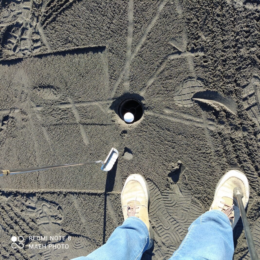 birds eye view of a golf hole with a golf ball in it. A close up of two feet and a putter. On the Nullarbor Links - some holes were grass, some sand and some oiled dirt, like this one.