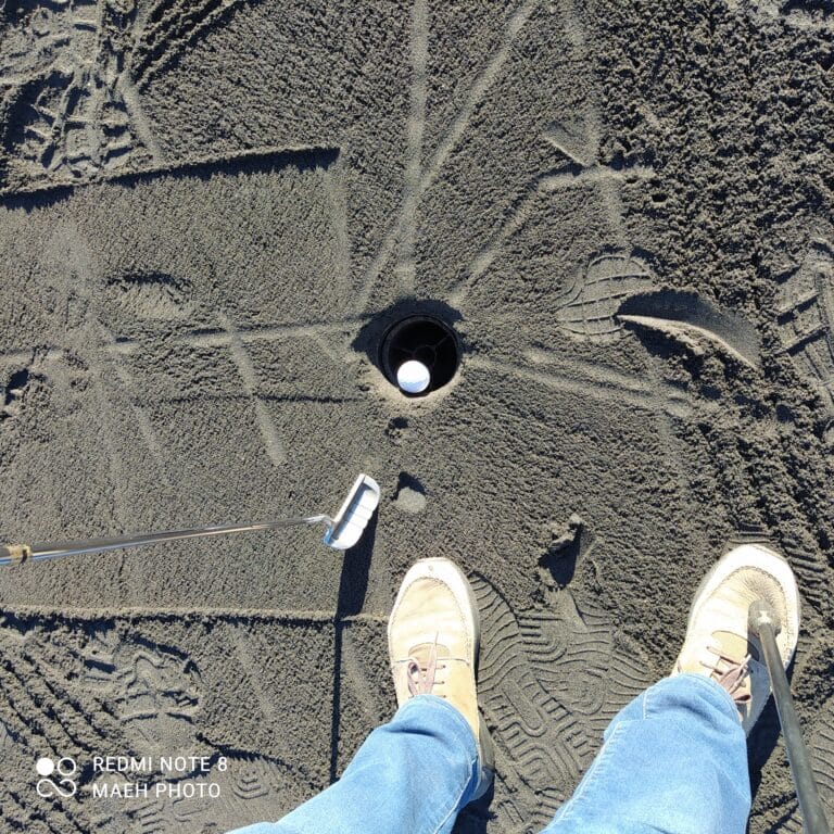 birds eye view of a golf hole with a golf ball in it. A close up of two feet and a putter. On the Nullarbor Links - some holes were grass, some sand and some oiled dirt, like this one.
