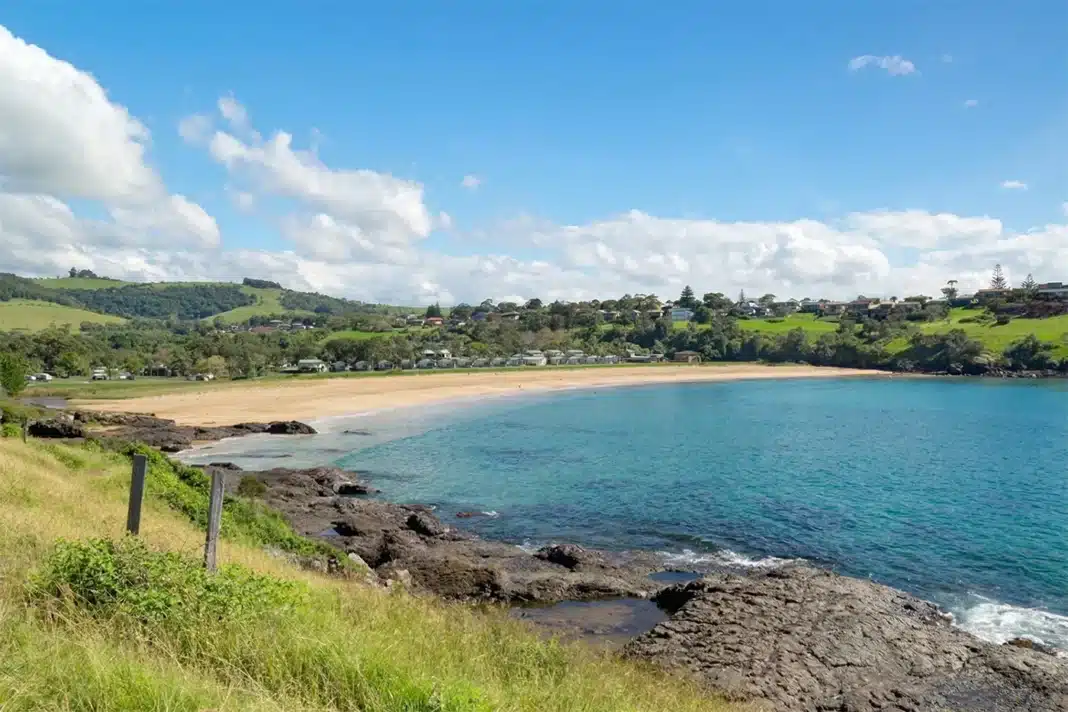 Sheltered sandy beach at Kiama NSW viewed from rocky foreshore, with crystal clear turquoise water, a holiday park and green rolling hills behind