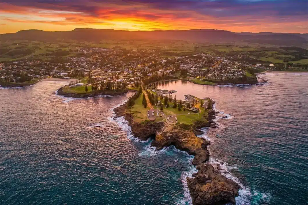 Aerial drone view of Kiama Harbour and Blowhole Point headland at sunset, with vivid orange and purple skies reflected on the ocean and the Illawarra escarpment in the background