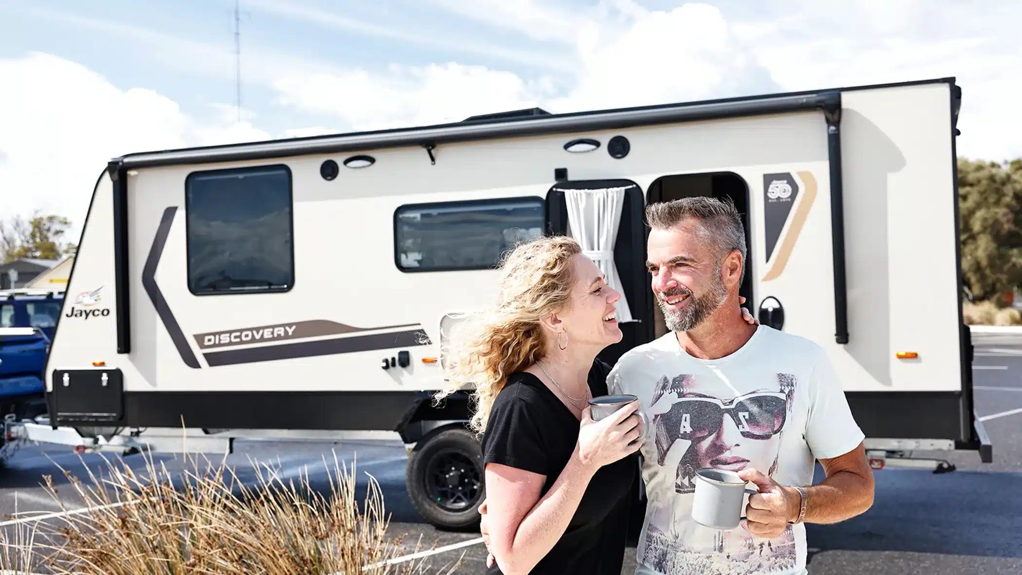 grey nomads in front of caravan with cup of coffee