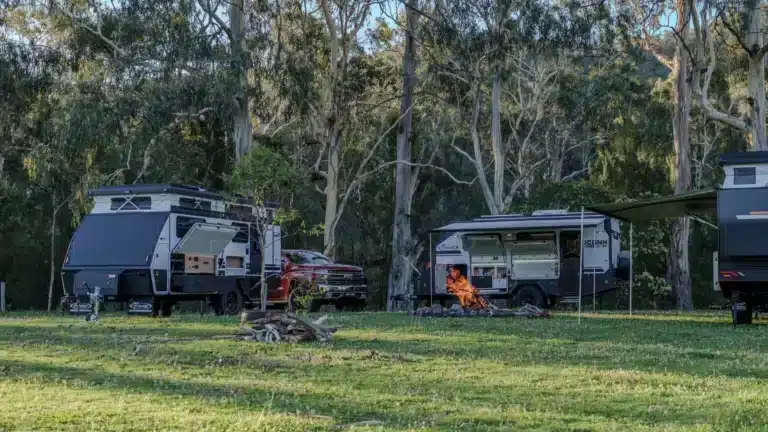 Off-road caravans and a ute set up on grass among gum trees, with a campfire burning between them