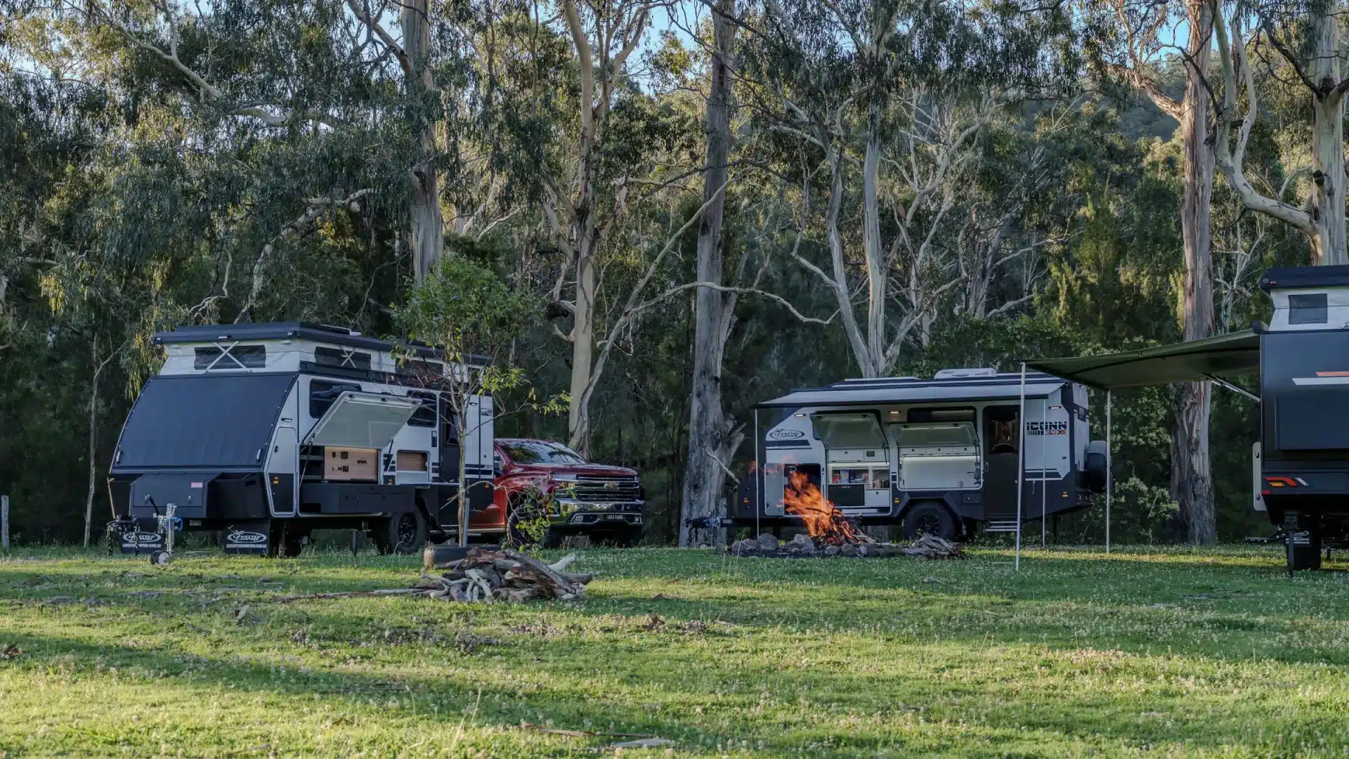 sunshine-coast-holiday-park-caravan-sites-demand Off-road caravans and a ute set up on grass among gum trees, with a campfire burning between them