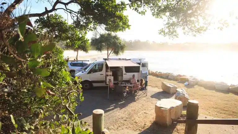 Campervan set up for free camping beside a coastal inlet with people sitting under an awning