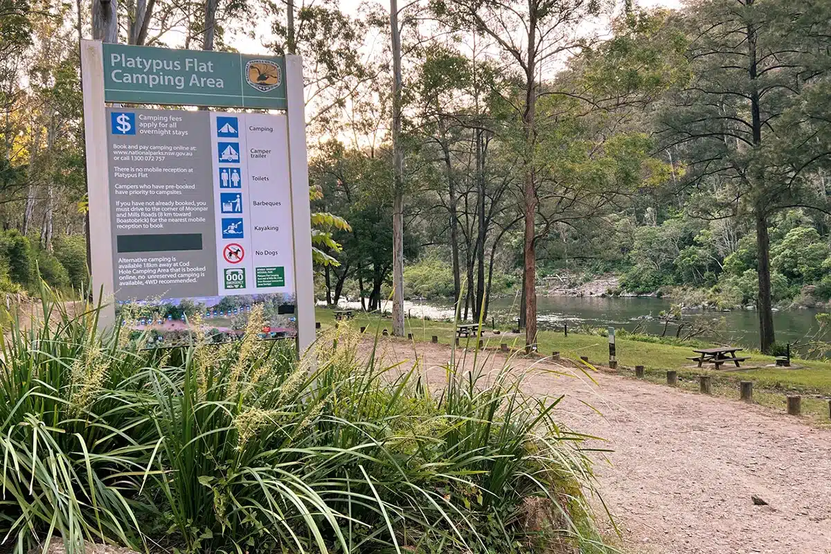 Sign for Platypus Flat Camping Area in New South Wales beside a riverside campground with picnic tables and trees.