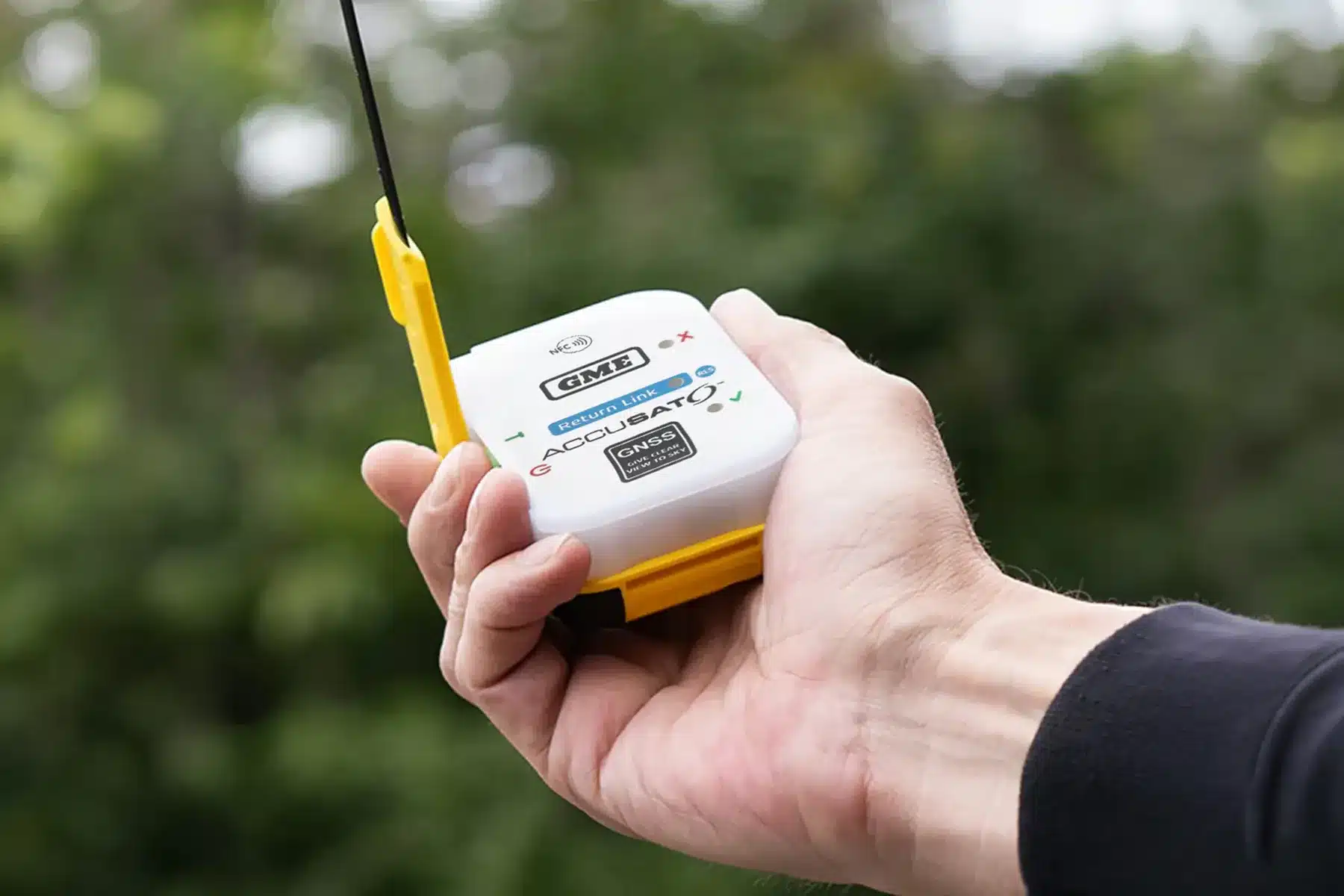 Hand holding an activated GME MT620GR personal locator beacon with antenna extended against a green bush backdrop
