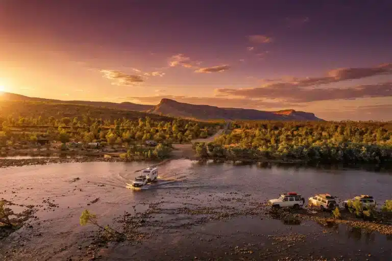 Caravan and 4WD crossing river through the Kimberley region of Western Australia with native trees in the background.