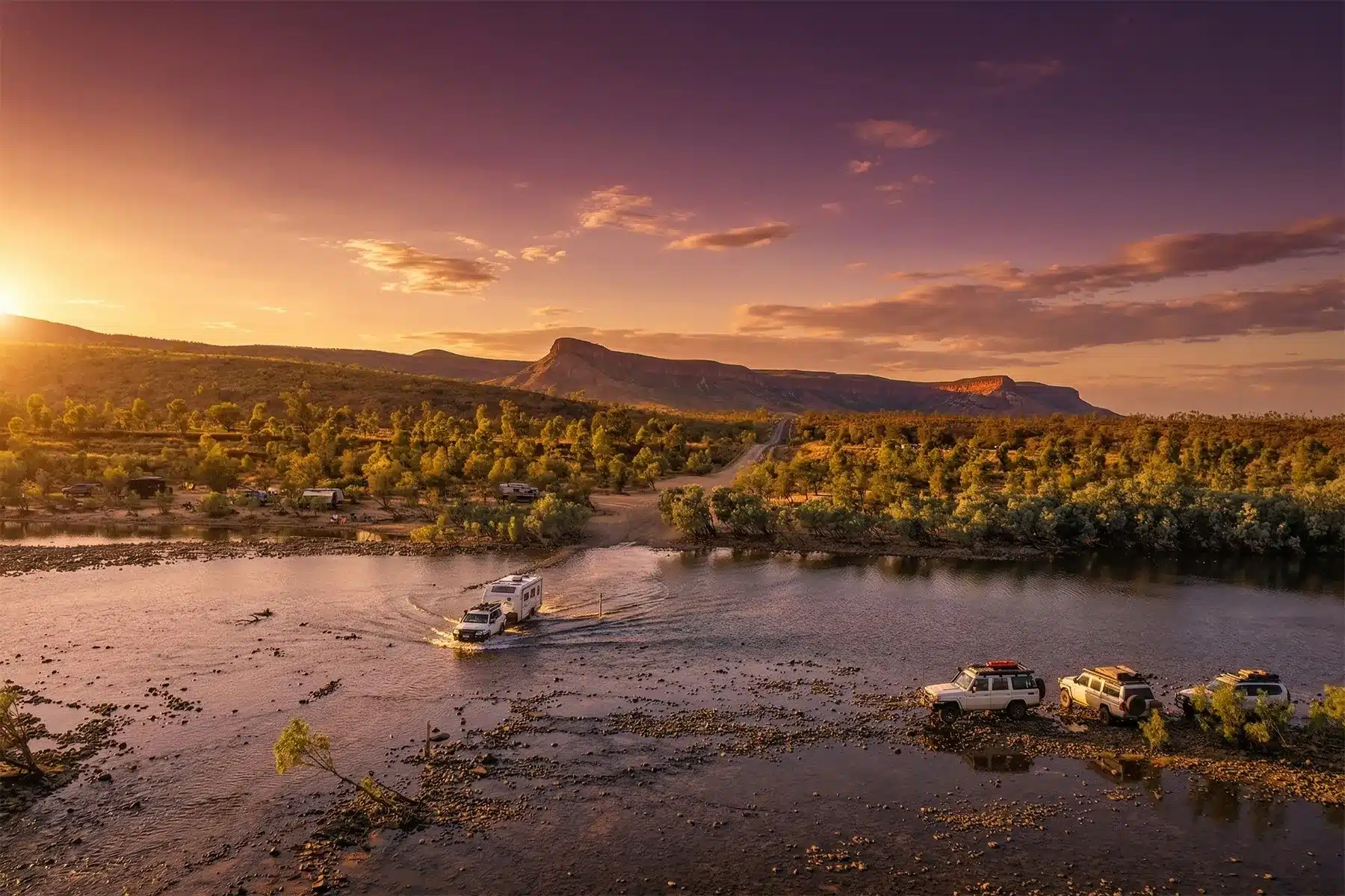 Caravan and 4WD crossing river through the Kimberley region of Western Australia with native trees in the background.