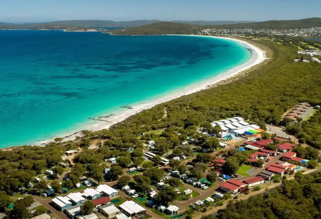 Drone view of a sprawling coastal caravan park situated right next to a pristine white sand beach and vibrant turquoise water.