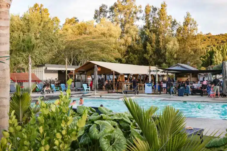 Holiday park guests relaxing by the pool and gathering at a lively outdoor entertainment pavilion and bar.