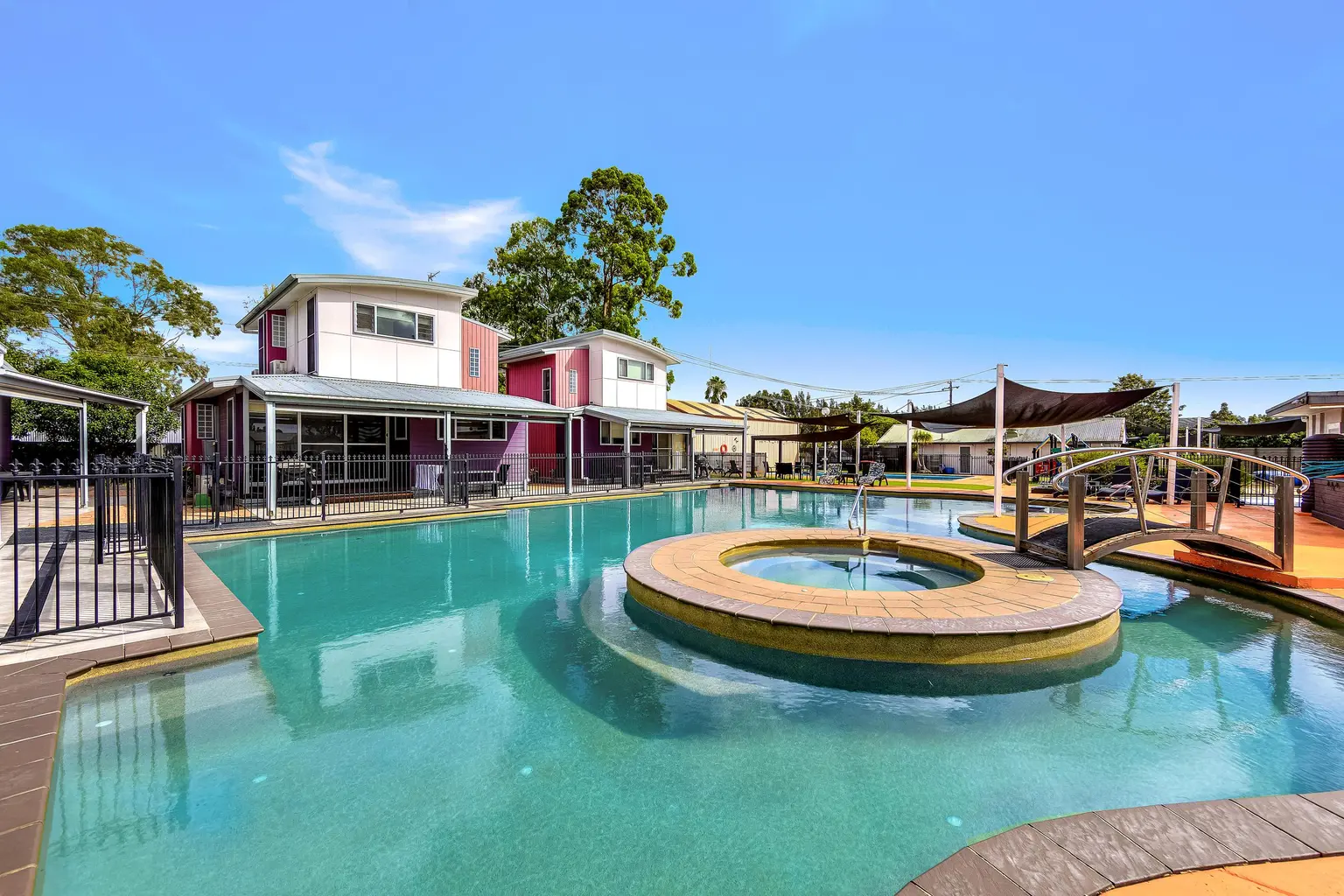 Bright blue holiday park swimming pool featuring a central circular spa connected by a small footbridge, with colourful cabins in the background.
