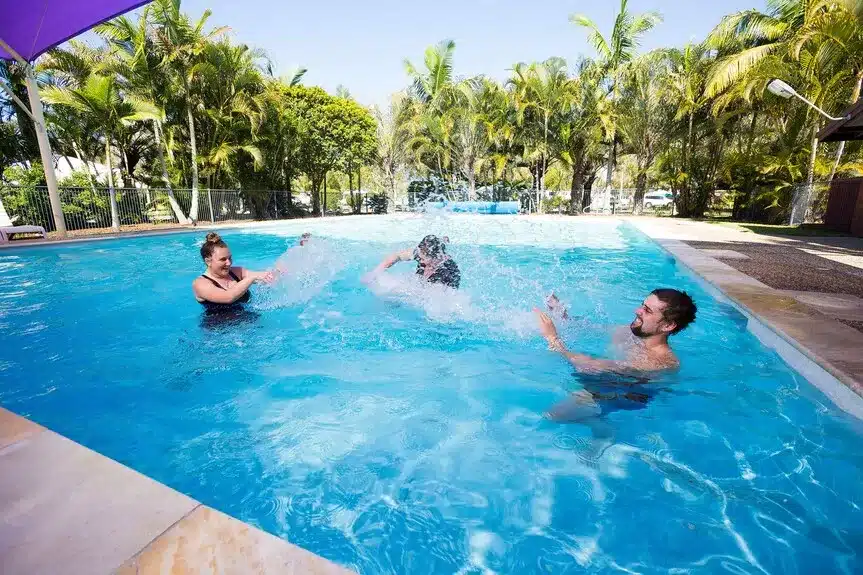 hree adults playfully splashing water in a sunlit, tropical holiday park swimming pool surrounded by tall palm trees.