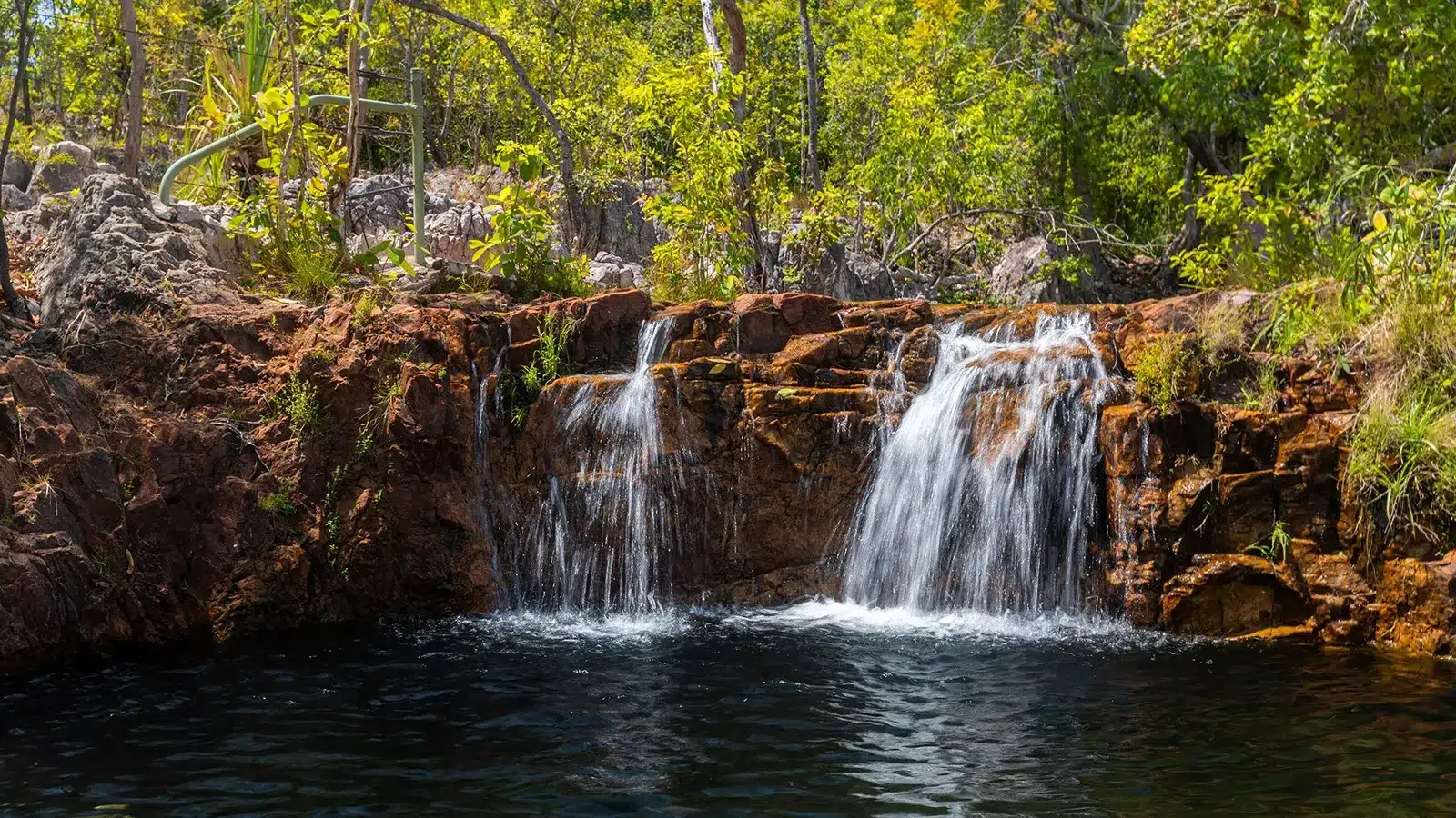 A natural waterfall cascading over rugged red rocks into a pristine swimming hole, typical of the day trips available in the Top End.
