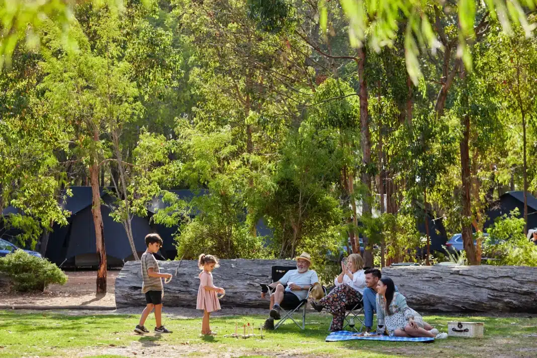 A multi-generational family relaxing with a picnic and playing lawn games outside luxury glamping tents set among tall eucalyptus trees at an Australian holiday park.