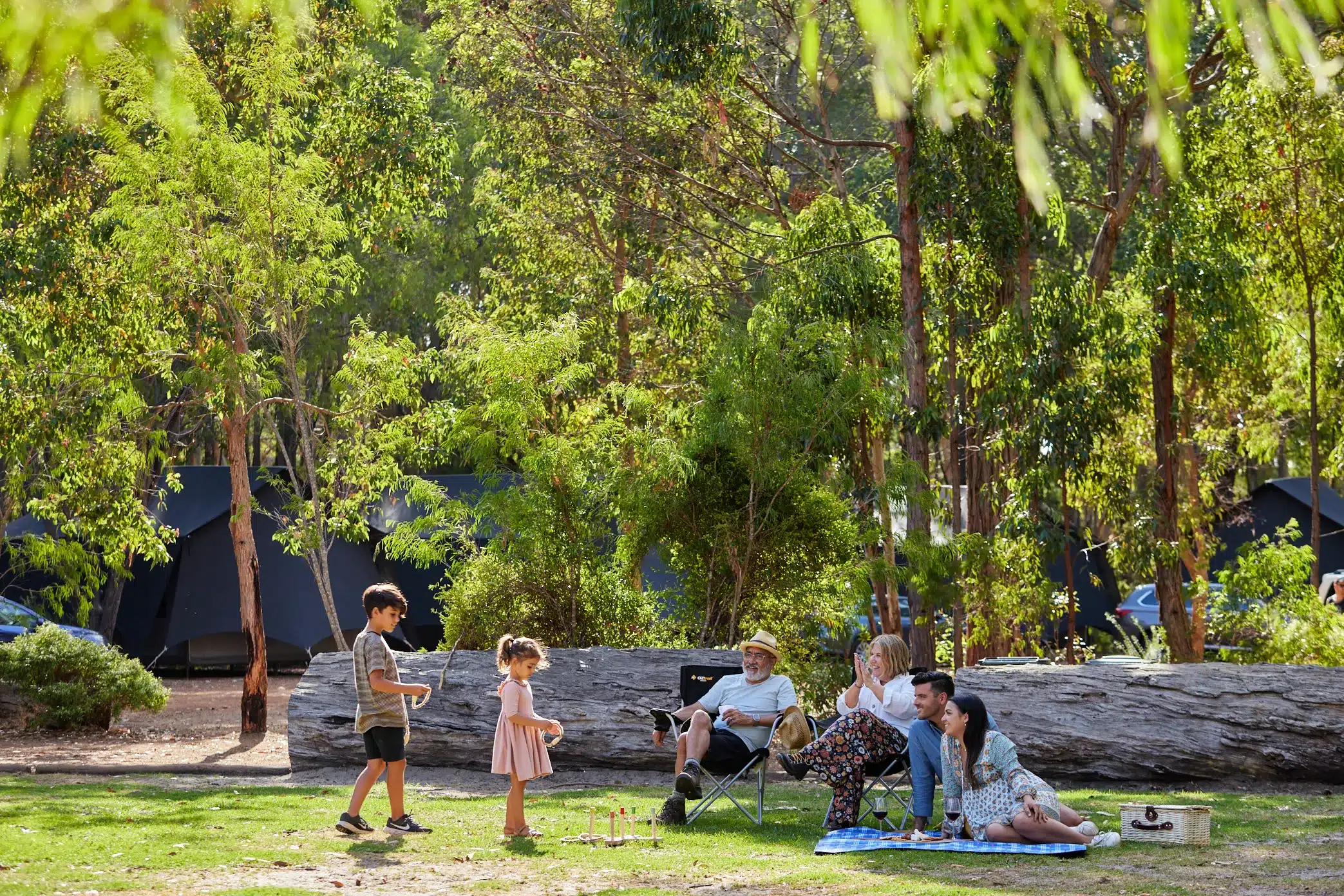 A multi-generational family relaxing with a picnic and playing lawn games outside luxury glamping tents set among tall eucalyptus trees at an Australian holiday park.