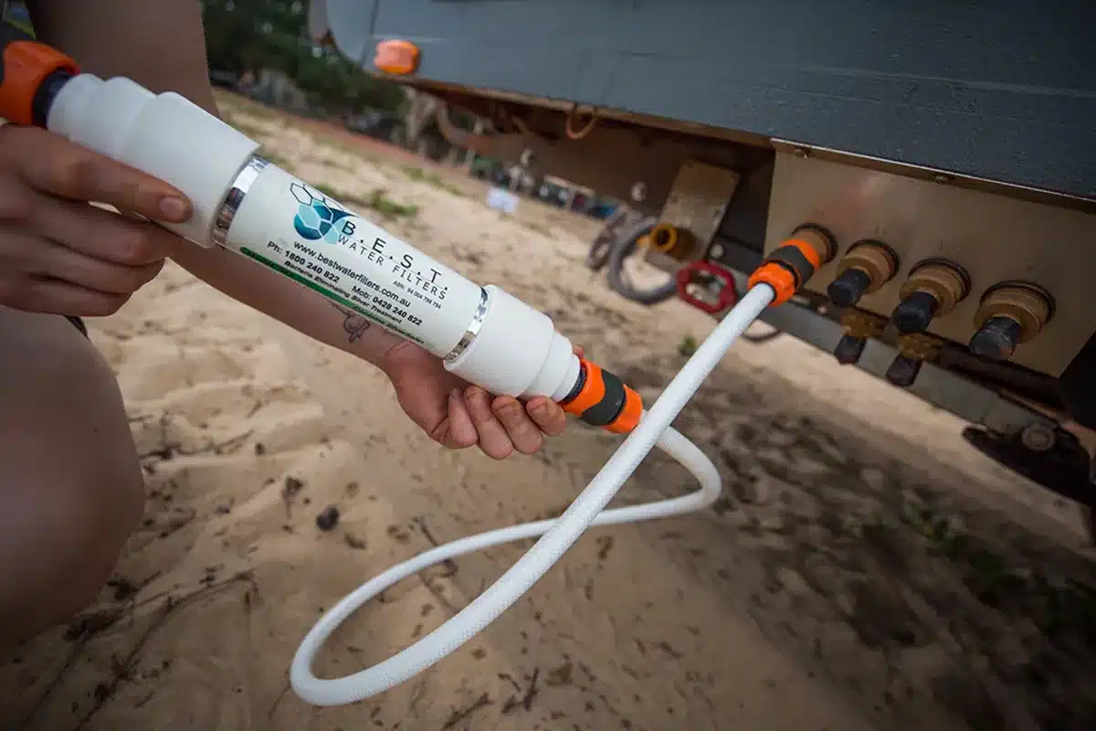 Hand holding a B.E.S.T. 322H inline water filter connected to a caravan water inlet via an orange click-on hose fitting