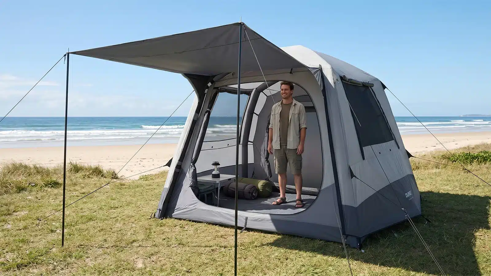 Man standing inside an open Dune 4WD Ultimate Air Tent on a grassy beachfront campsite, demonstrating the tent's full stand-up interior height