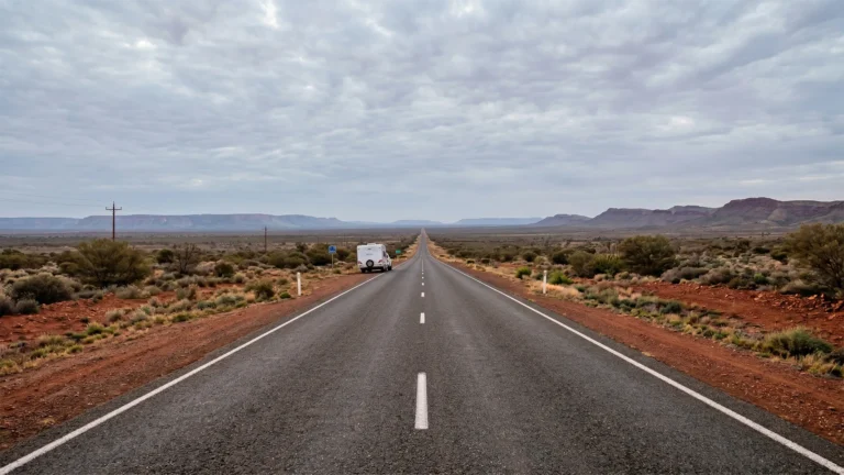 A white caravan travelling along a long straight outback highway in Central Australia, with red dirt shoulders, low scrubland and flat-topped ranges stretching to the horizon under an overcast sky