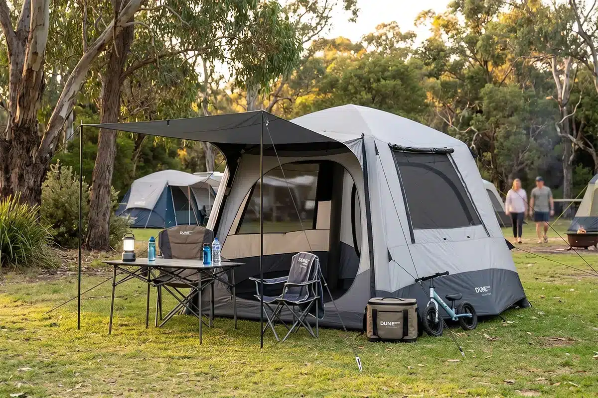Dune 4WD Ultimate Air Tent set up at a bush campground at dusk, with a camp table, lantern, chair, cooler bag, and kids' balance bike outside, and two people in the background