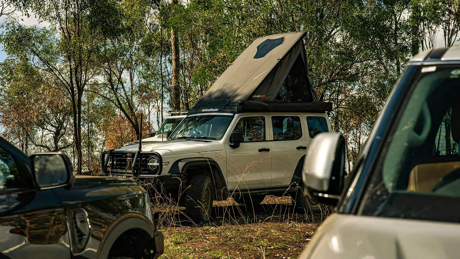 Hard shell rooftop tent deployed on a white 4WD wagon in Australian bush, clamshell lid open showing sleeping platform, gum trees and dappled light in background
