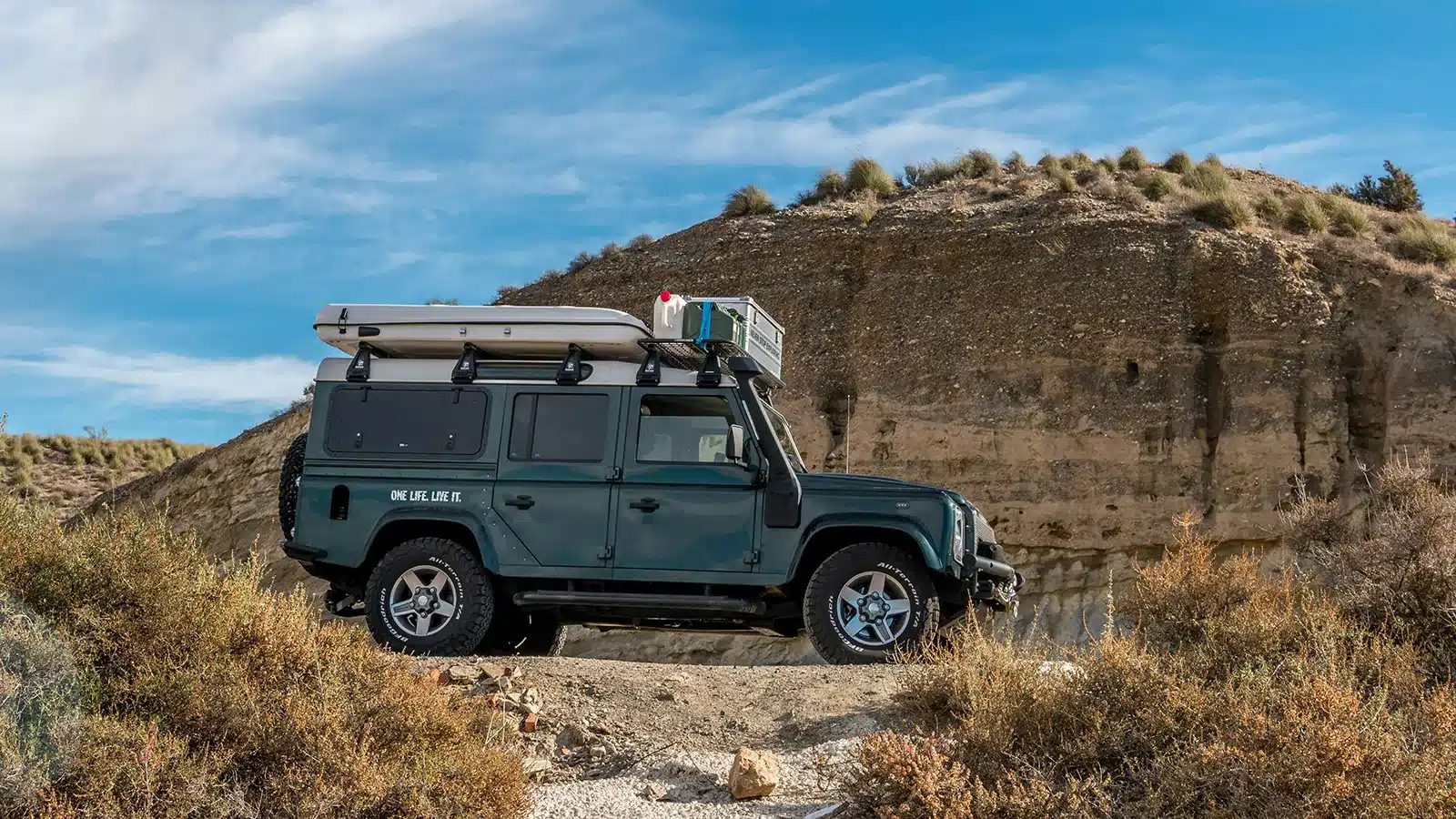 Land Rover Defender with hard shell rooftop tent packed flat on roof racks, closed and low-profile on a rocky outback track, brown cliff face and dry scrub behind
