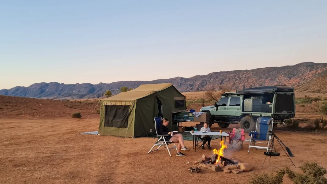Mother and child sitting around a campfire at a remote Australian outback private campsite with a canvas tent and 4WD against red rocky ranges