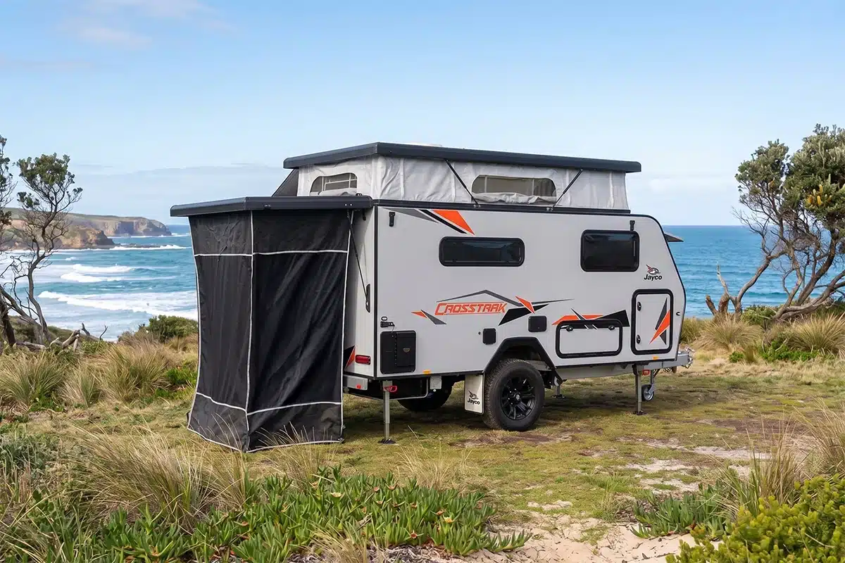 Jayco CrossTrak 13ft hybrid pop-top caravan set up on a coastal headland with pop-top raised and awning privacy screen deployed, Southern Ocean visible in the background