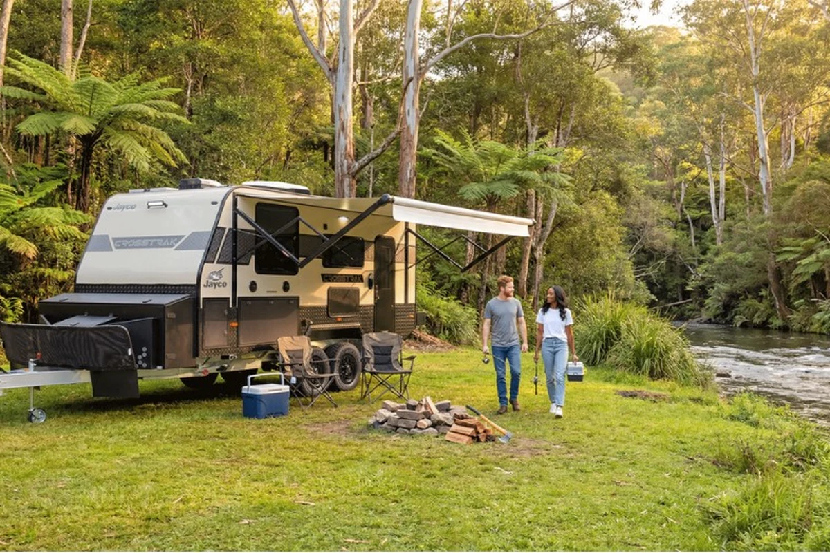 Jayco CrossTrak hybrid caravan set up at a riverside bush campsite with awning deployed, couple walking toward campfire in Australian rainforest setting