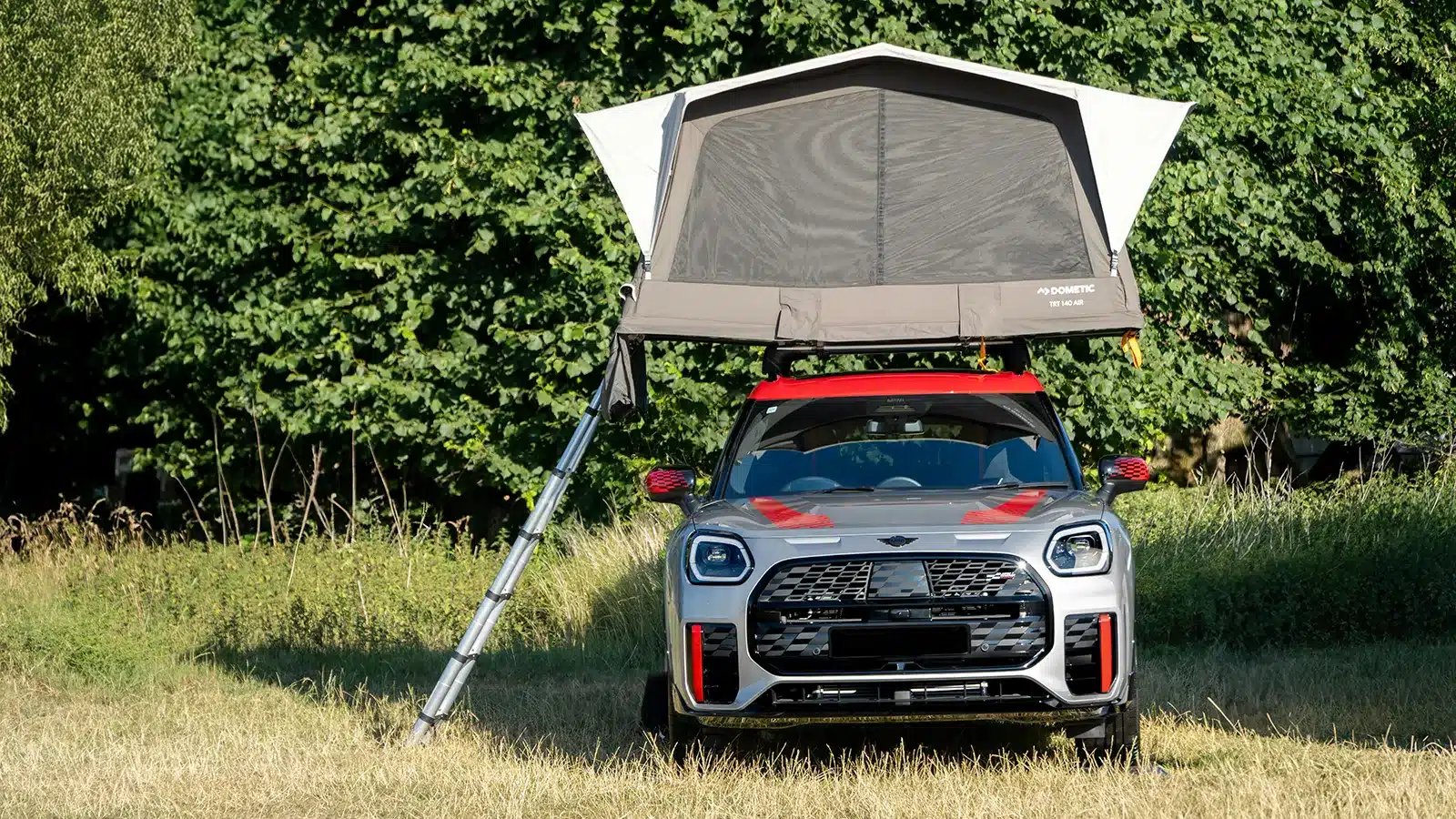 Soft shell rooftop tent fully deployed on a compact SUV in a green field, A-frame canvas raised with mesh walls open, tall trees in background