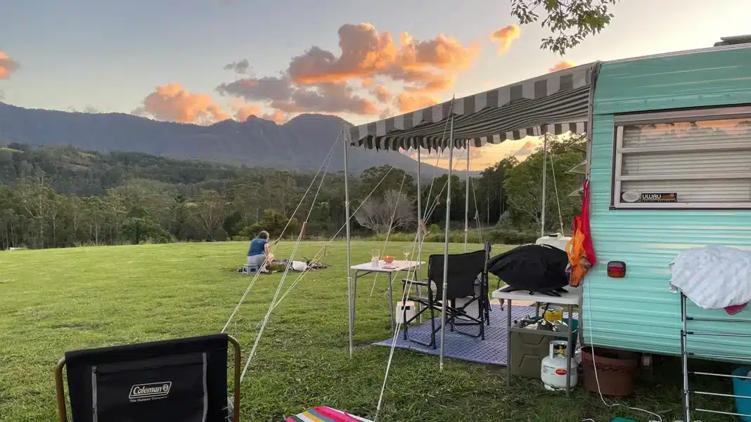 Vintage mint-green caravan with striped awning set up at a private property campsite, with lush green paddock, mountain ranges and vivid sunset clouds in the background