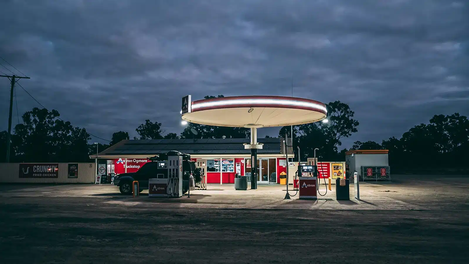 A remote Australian roadside service station lit up at dusk, with a 4WD parked at the bowser under a glowing oval canopy, surrounded by dark scrubland and overcast skies