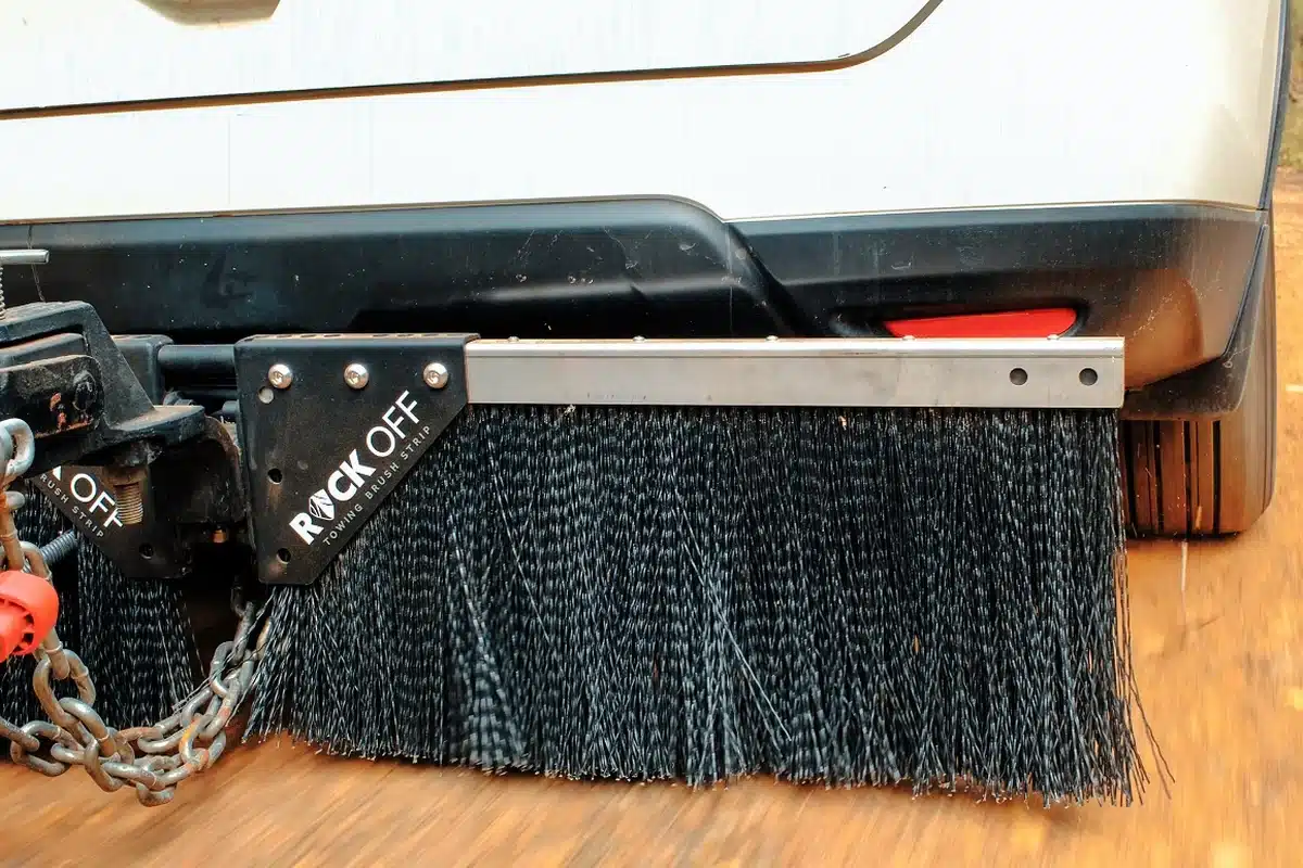 Rock Off towing brush strip stone guard mounted under a tow vehicle near the hitch, showing dense polypropylene bristles and aluminium extrusion bar, vehicle in motion on a red dirt road