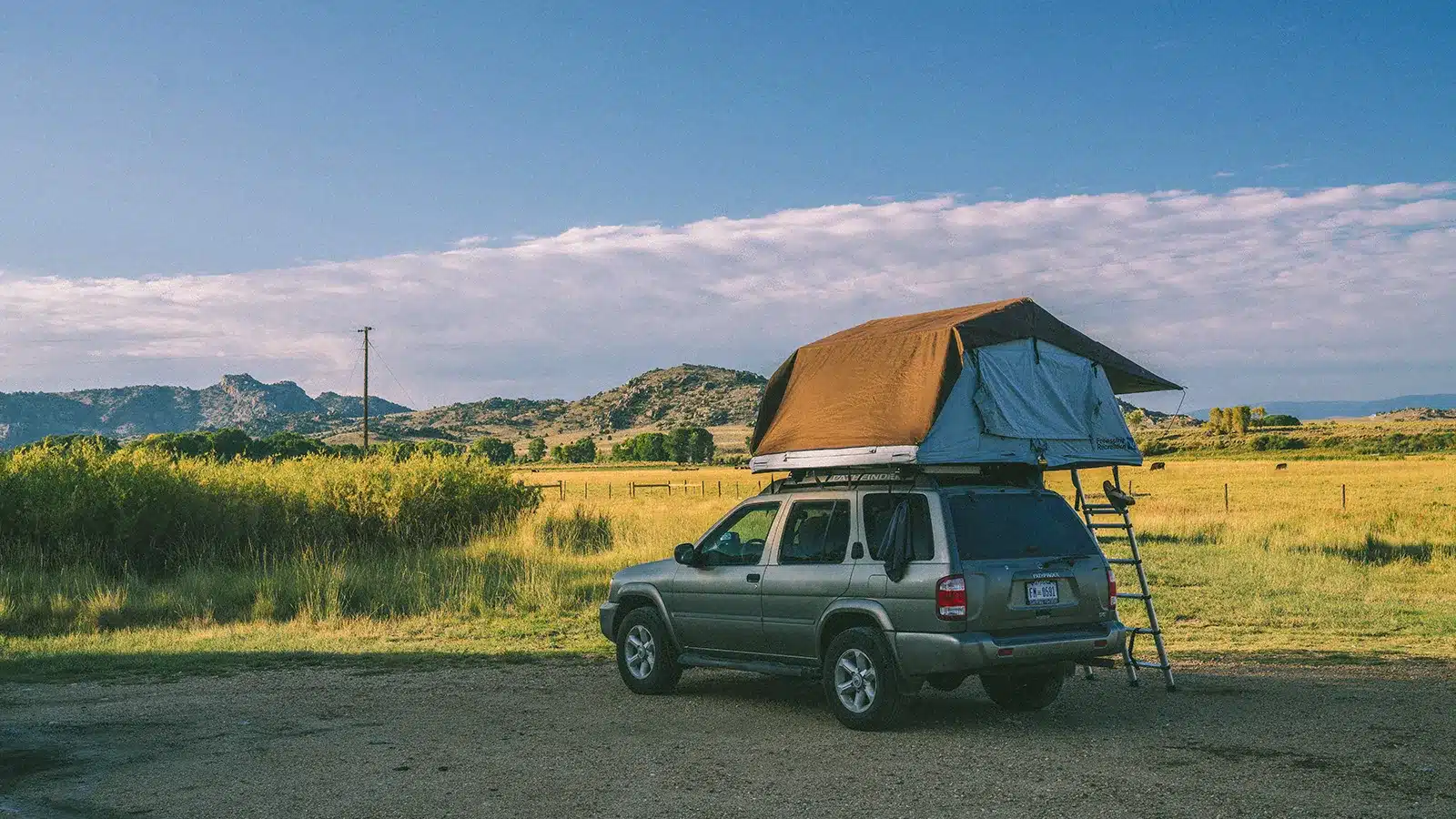 Soft shell rooftop tent open on a silver SUV parked in open countryside, orange canvas fly extended with ladder deployed, wide rural landscape under blue sky