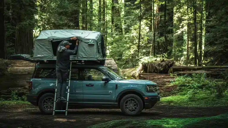 Person climbing ladder to access a hard shell rooftop tent on a teal Ford Bronco Sport, surrounded by tall redwood forest, tent partially open showing setup process