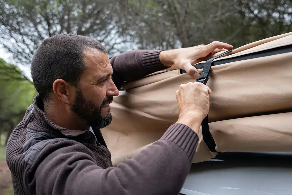 Man strapping down tan canvas on a soft shell rooftop tent ready for pack-down, hands tightening webbing straps over folded canvas on roof bars, overcast bush setting