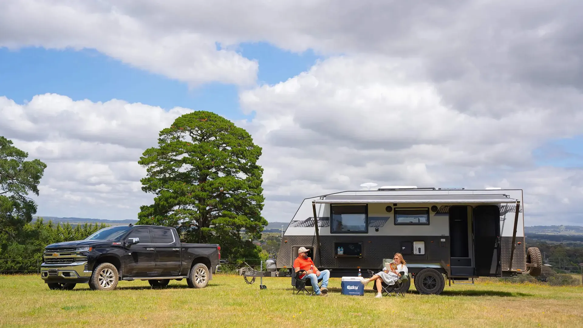 Couple relaxing beside an Australian-built off-road caravan and Chevrolet Silverado tow vehicle in a country paddock