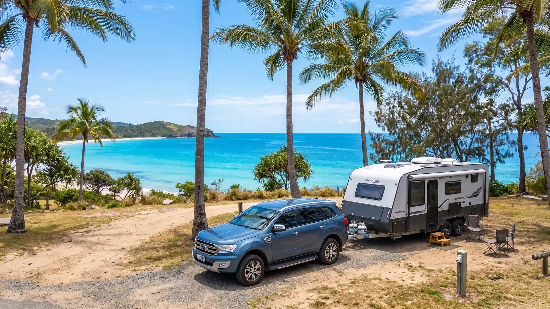 Blue SUV towing a caravan at a coastal campsite in Queensland, with palm trees framing a turquoise ocean bay and headland in the background.
