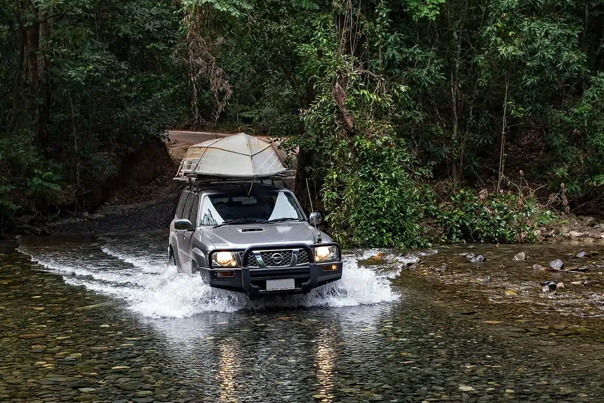 A silver four-wheel-drive vehicle with a small metal boat strapped to its roof rack splashes through a shallow, rocky river crossing surrounded by dense, green tropical rainforest.