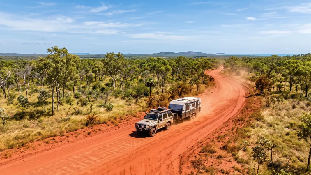 cape-york-caravanning An aerial view of a 4x4 vehicle towing a caravan, kicking up dust as it drives along a winding, red dirt outback road through a green, sparsely wooded landscape with ocean visible in the far distance.