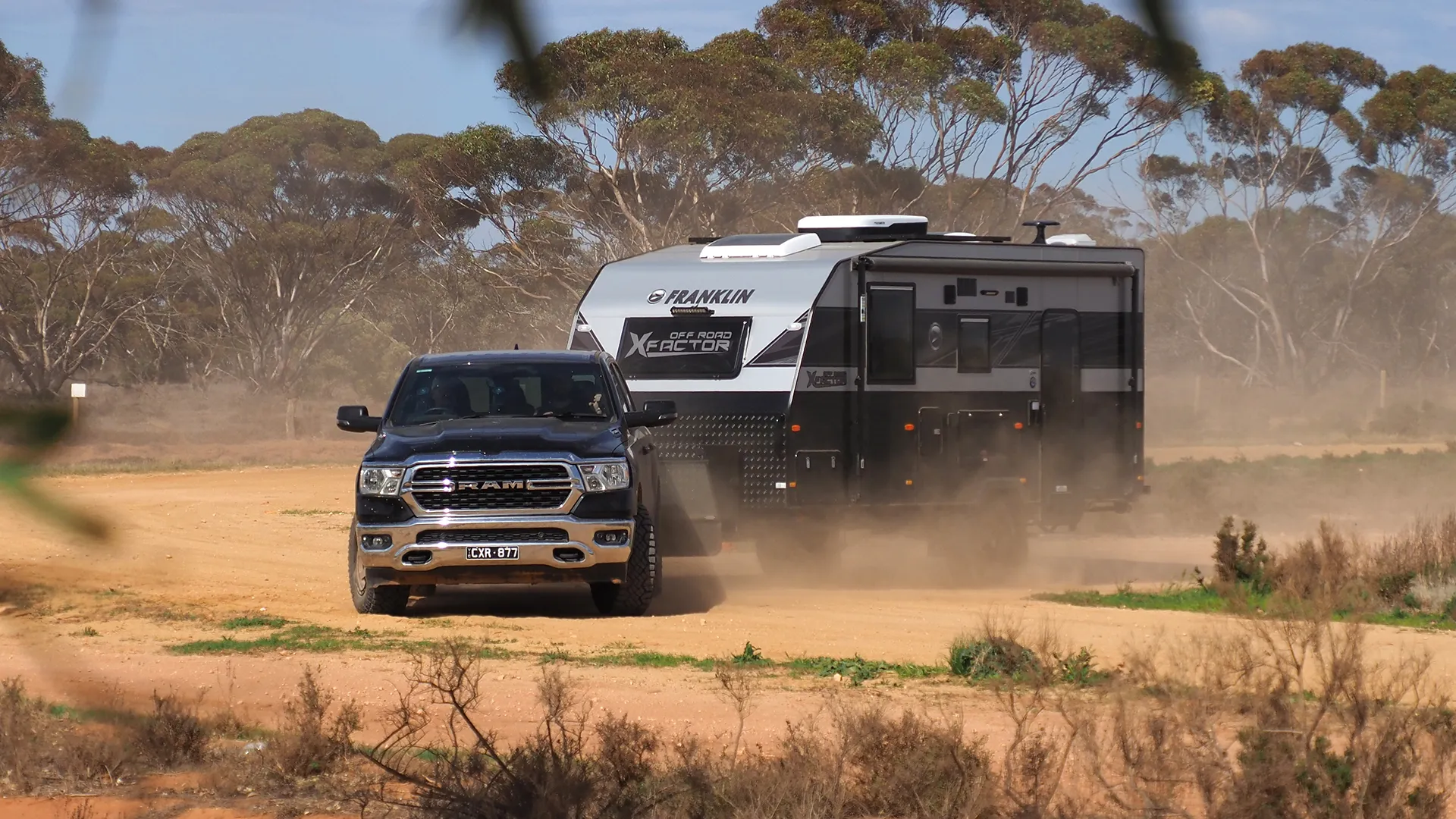 RAM ute towing a Franklin Off Road X Factor caravan along a dusty gravel road lined with mallee scrub