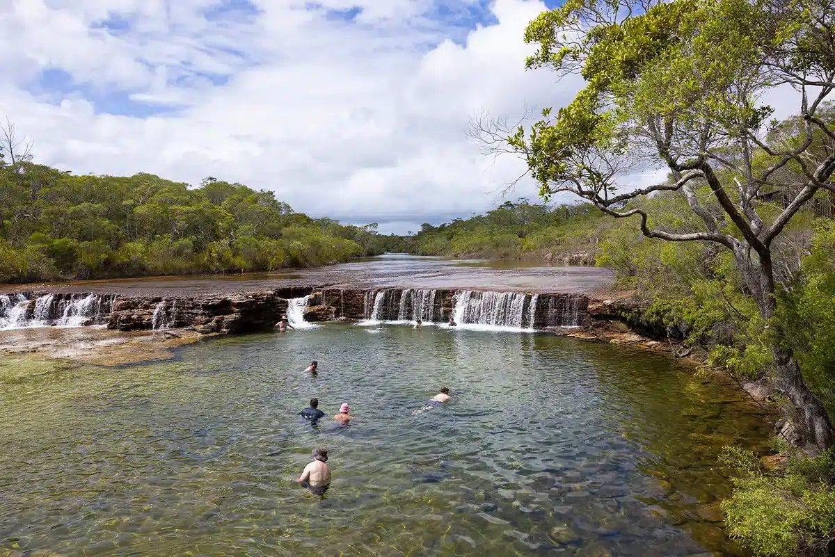 Several people swimming and wading in a clear, shallow natural pool at the base of a wide, low waterfall, surrounded by dense green trees under a bright sky with fluffy white clouds.