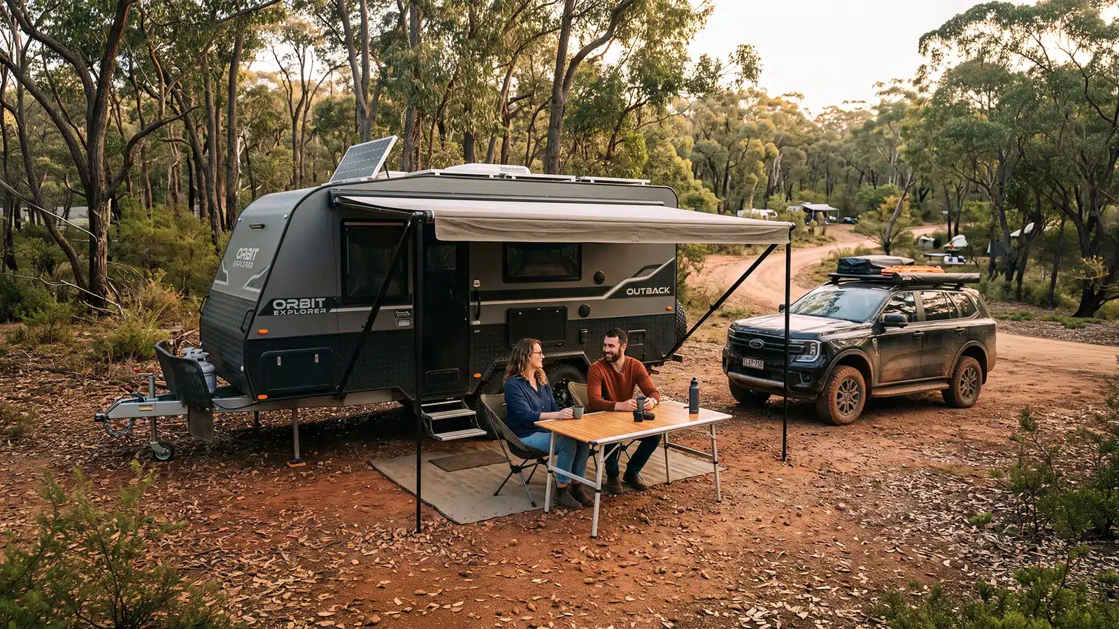 A young, casual-clothed couple smiles while sitting at a portable table under the awning of a modern 'Orbit Explorer Outback' caravan, with a dark Ford Everest 4x4 SUV parked nearby in a wooded Australian campsite.