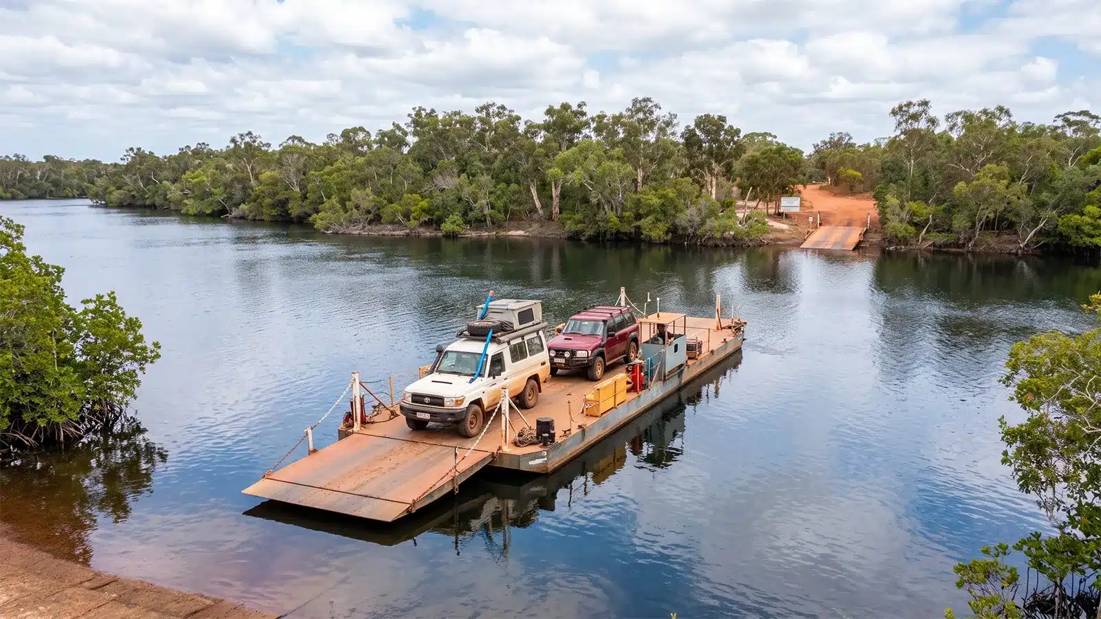A flat, cable-operated ferry transporting a white 4x4 and a maroon 4x4 across a wide, calm, dark-blue river, with dense green forest lining both banks and a dirt boat ramp visible on the far side.
