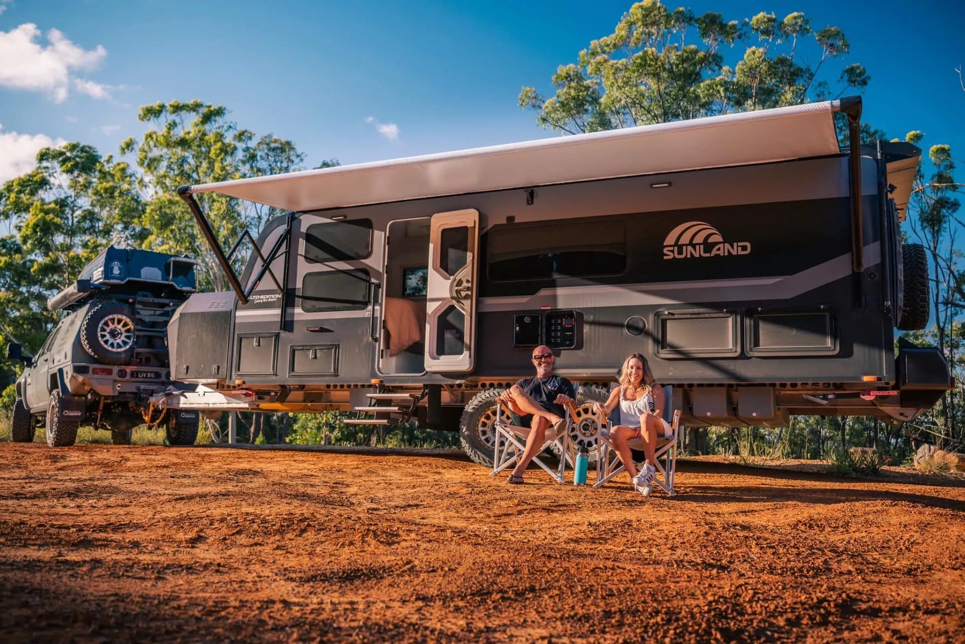 An adult couple sits comfortably in portable chairs under the wide awning of a premium, large-format, dark grey 'Sunland' off-road caravan on red dirt, with their massive dual-cab towing ute parked nearby.