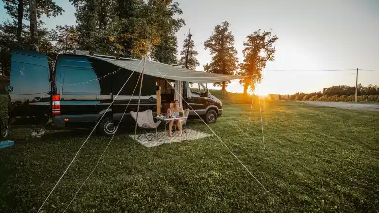 Black Mercedes Sprinter campervan with a shade awning set up on grass at sunset, camp chairs and table underneath