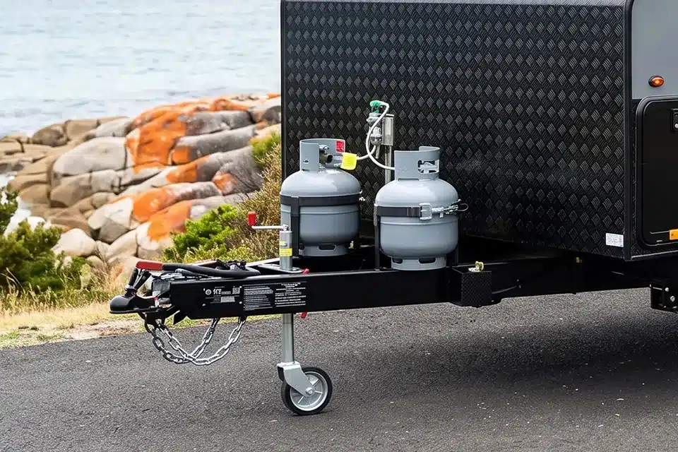Two grey LPG gas bottles mounted on a caravan A-frame with a changeover regulator, parked beside a rocky coastline