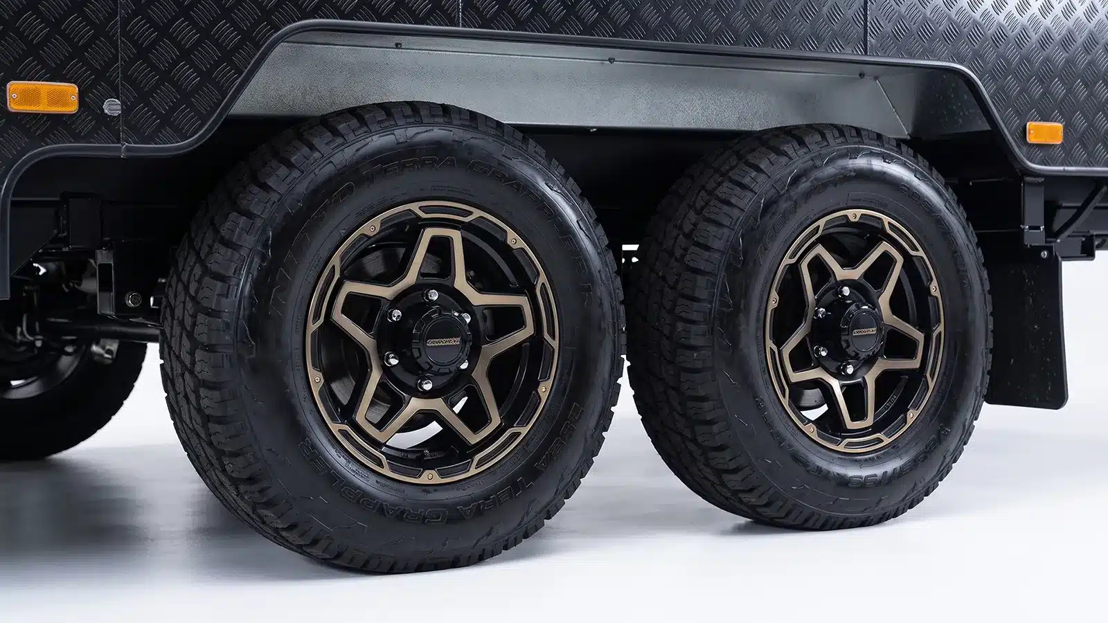 Tandem axle caravan wheels with all-terrain tyres and bronze alloy rims below a checkerplate body