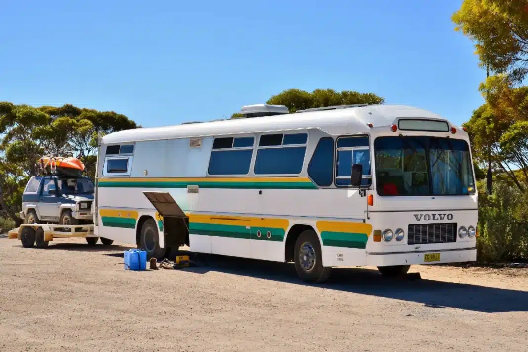 White converted Volvo coach with a four-wheel drive on a trailer behind it, parked among mallee trees at Eucla