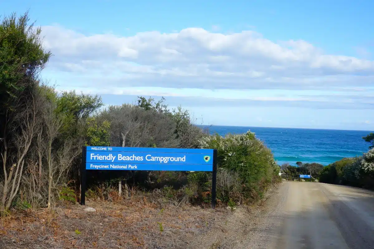 Friendly Beaches Campground sign in Freycinet National Park, Tasmania, beside a gravel road leading toward the ocean